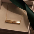 Gold necklace with a bar pendant on a green ribbon inside a box.