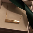 Gold necklace with a bar pendant on a green ribbon inside a box.